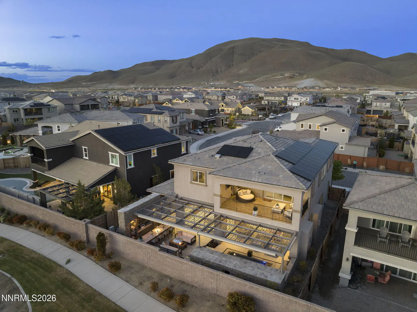 Aerial view of a two-story house with solar panels, a covered patio, and a balcony, in a neighborhood with a mountain backdrop.
