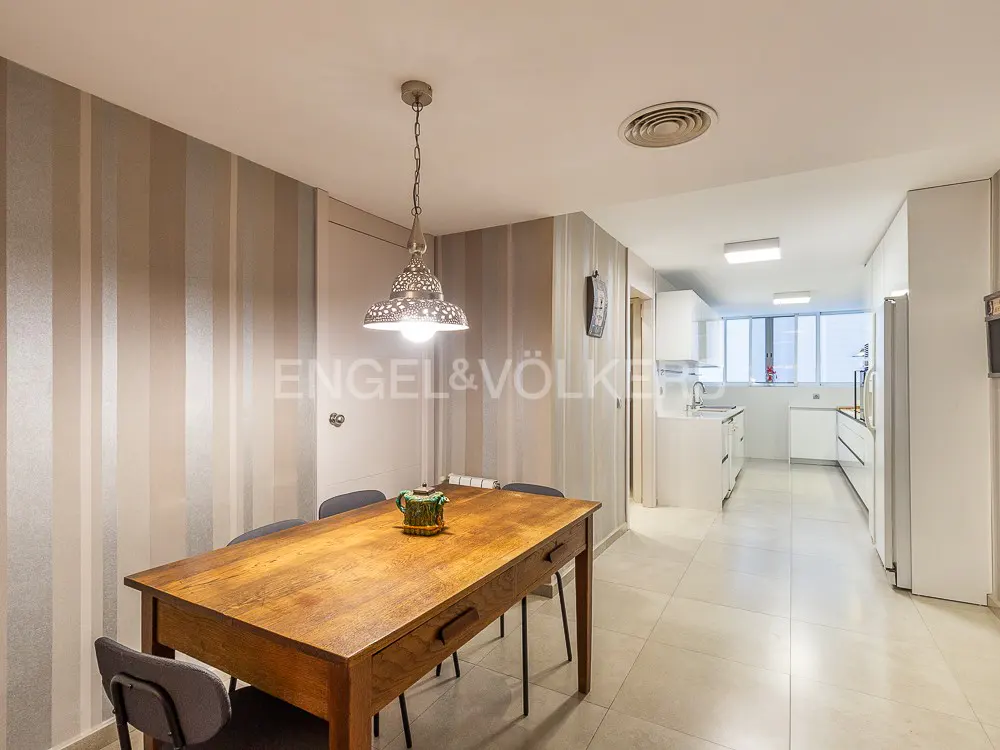 Dining area with a wooden table, gray chairs, and a decorative teapot. Striped wallpaper and a view into a white kitchen.