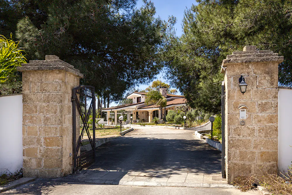 Gated entrance to a two-story house with a brown roof, seen through stone pillars and trees under a blue sky.
