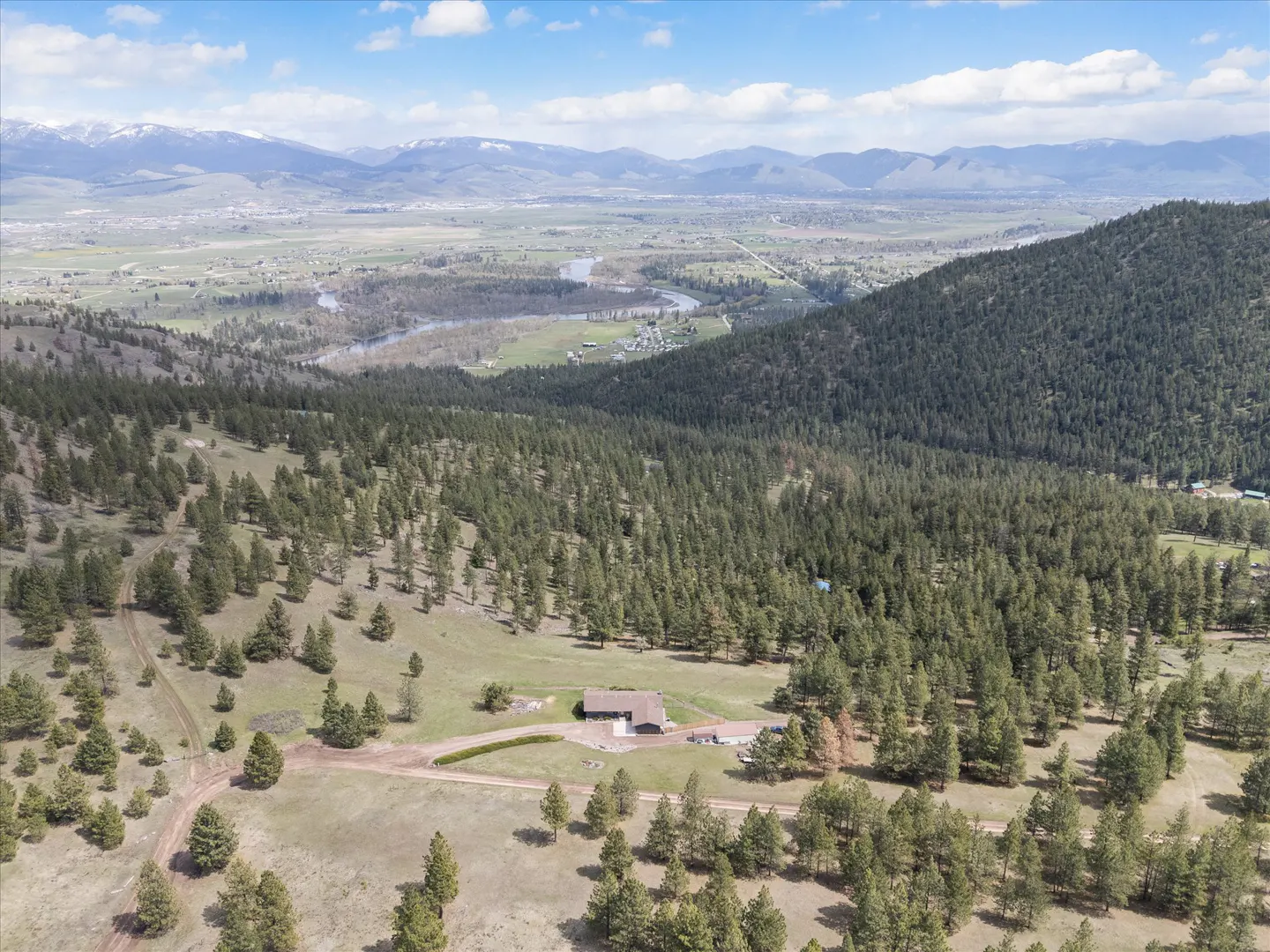 Aerial view of a brown house nestled in a green, pine-covered hillside with mountains in the background under a blue, cloudy sky.