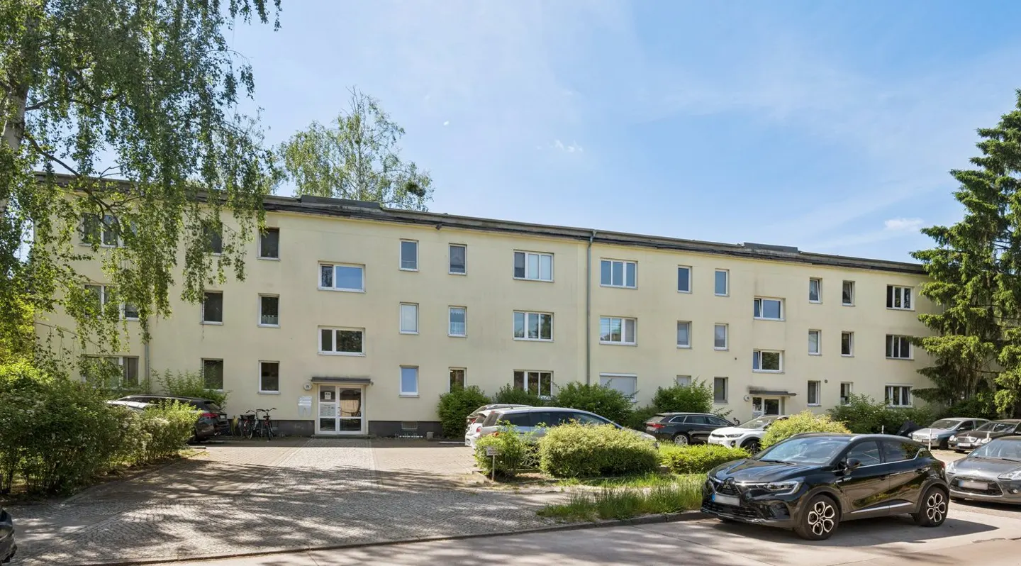 A three-story, light-yellow apartment building with a paved parking area in front, surrounded by trees and parked cars.