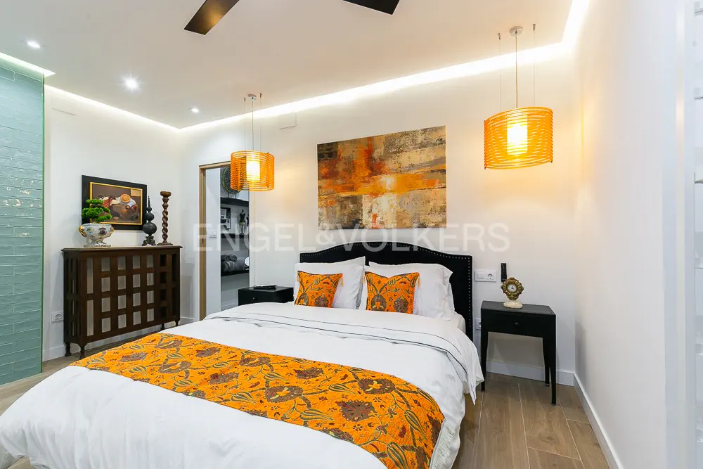 Modern bedroom with a white bed, orange patterned runner and pillows, two orange pendant lights, and abstract wall art.