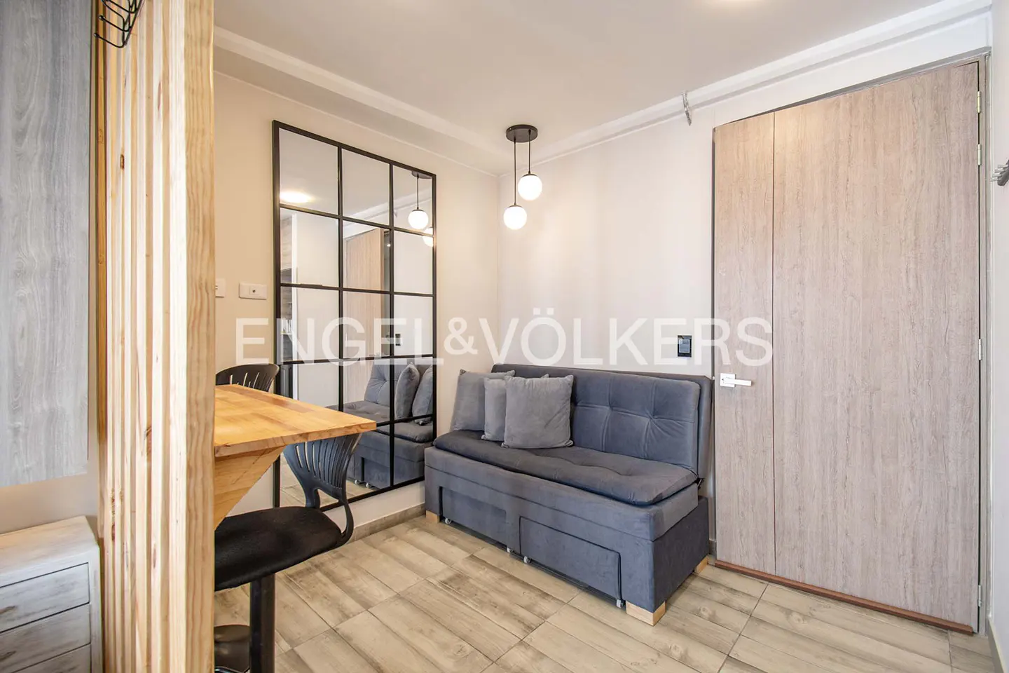 A compact living space with a gray sofa, wooden table, and a black-framed mirror. A light wood door is on the right.