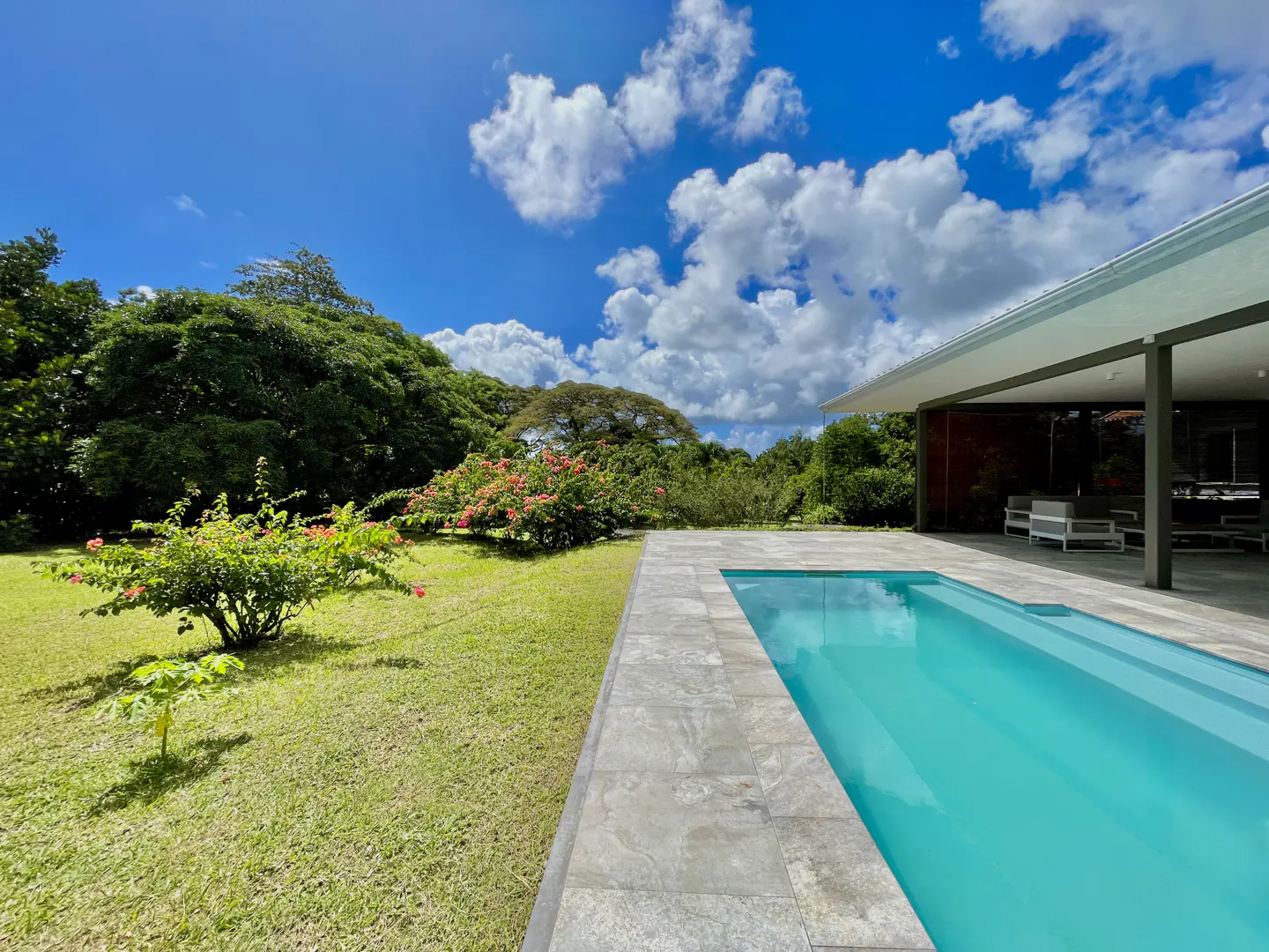 A turquoise pool is surrounded by a gray stone patio, green grass, and lush trees under a blue sky with white clouds.