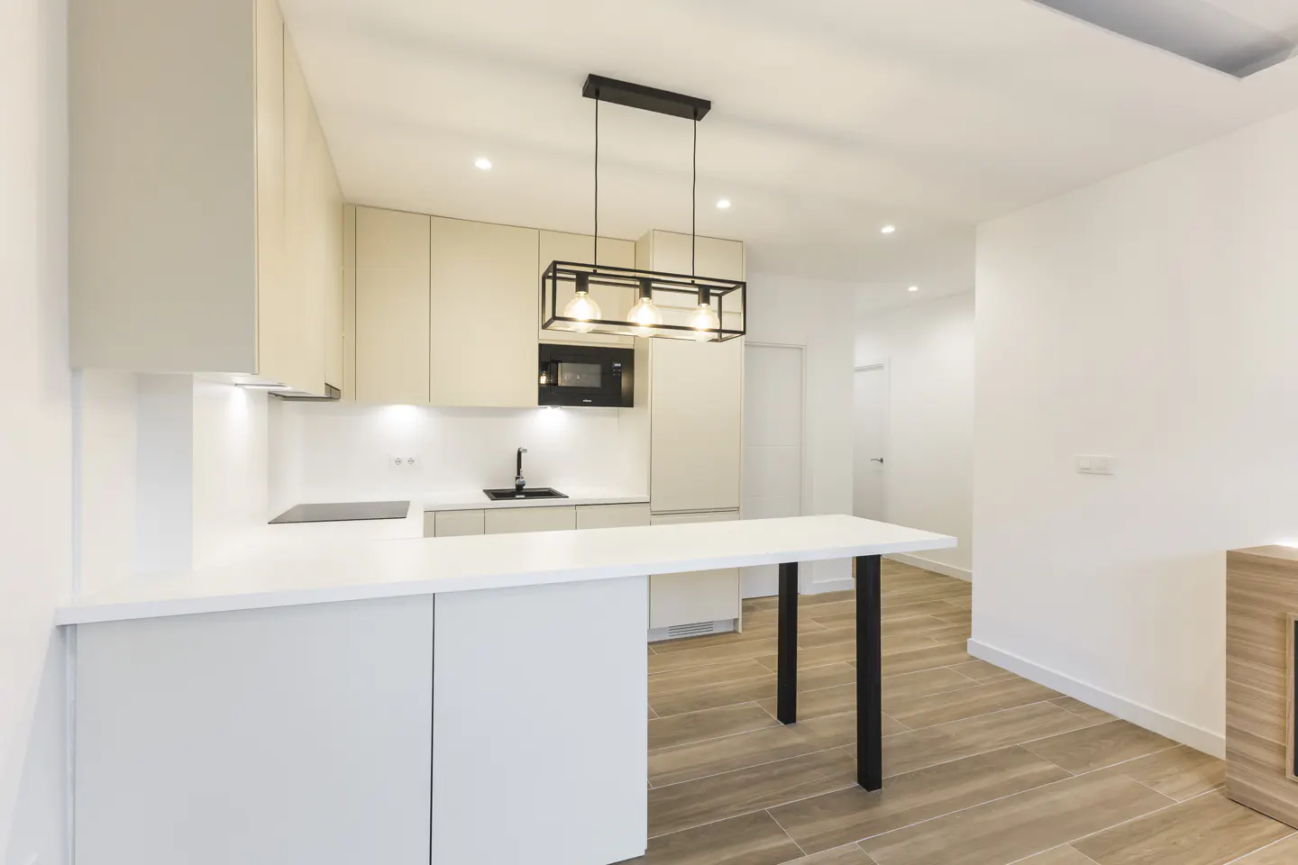 Bright, modern kitchen with white cabinets, countertops, and island. Black accents include a sink, microwave, and pendant light fixture. Wood-look tile flooring.