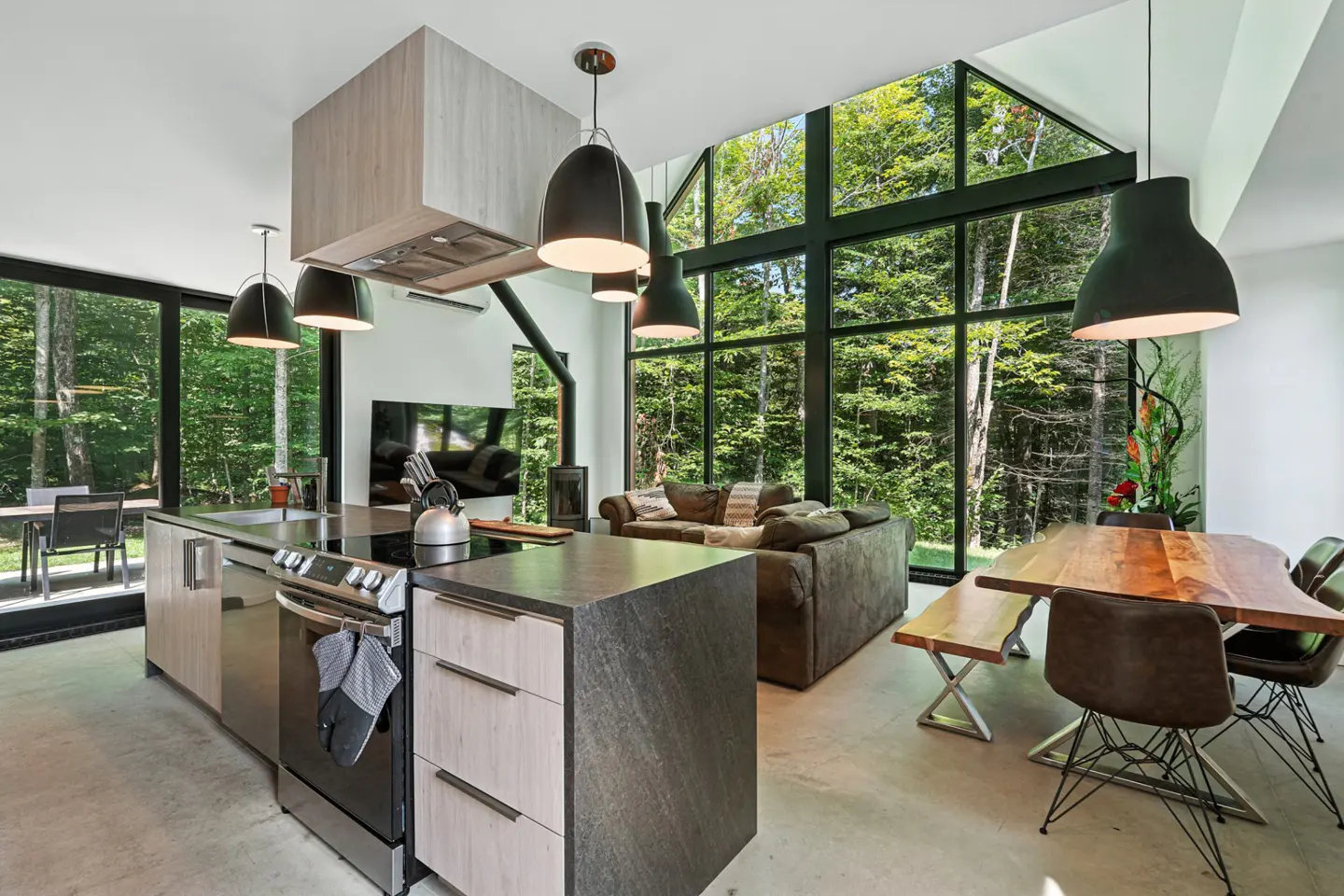 Open-concept kitchen and living room with large windows overlooking a forest. Kitchen island, dining table, and sofa are visible.