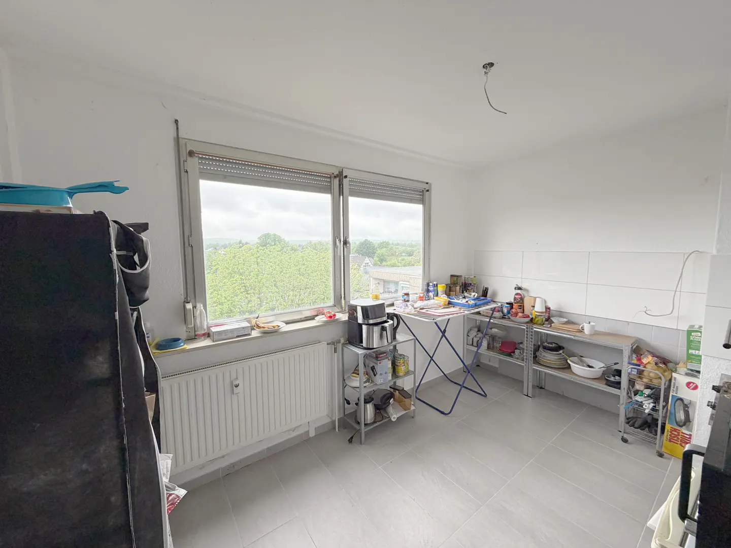Bright kitchen with gray tile floor, white walls, and a large window overlooking greenery. Shelves hold various kitchen items.