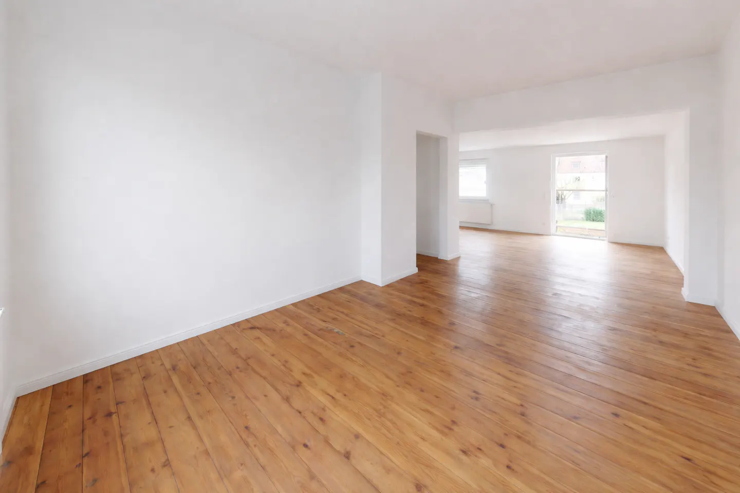 Empty room with light brown wood floors and white walls. A doorway leads to another room with a window.