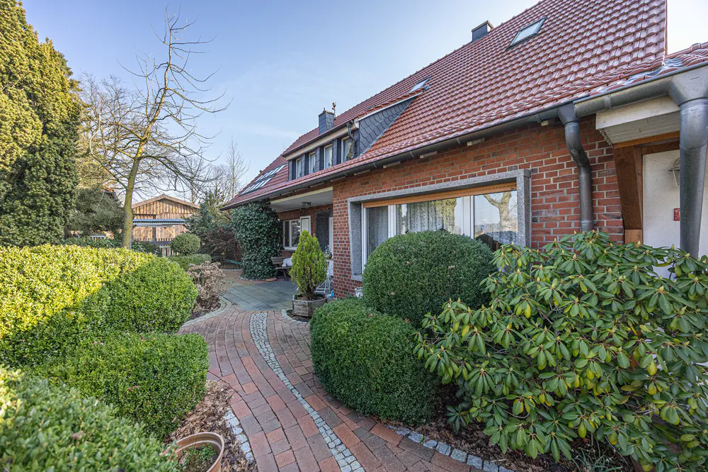 Exterior of a brick house with a red tile roof and a brick walkway surrounded by green bushes.