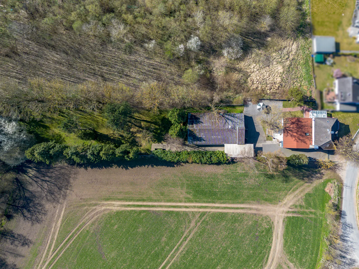 Aerial view of a property with a red-roofed house, barn, green field with tire tracks, and trees.