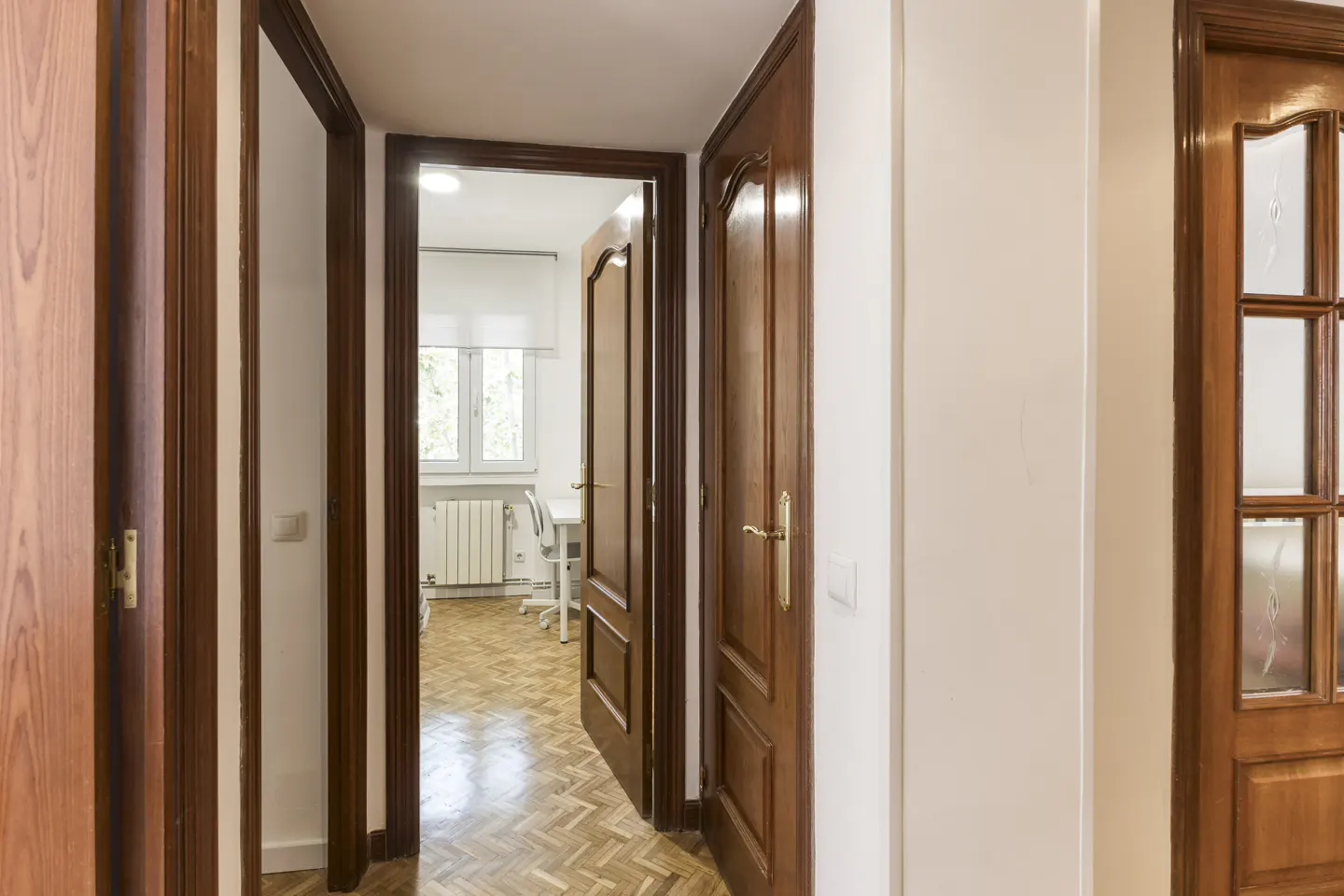 Hallway with wooden doors and herringbone floor. A room with a window, desk, and radiator is visible through an open doorway.