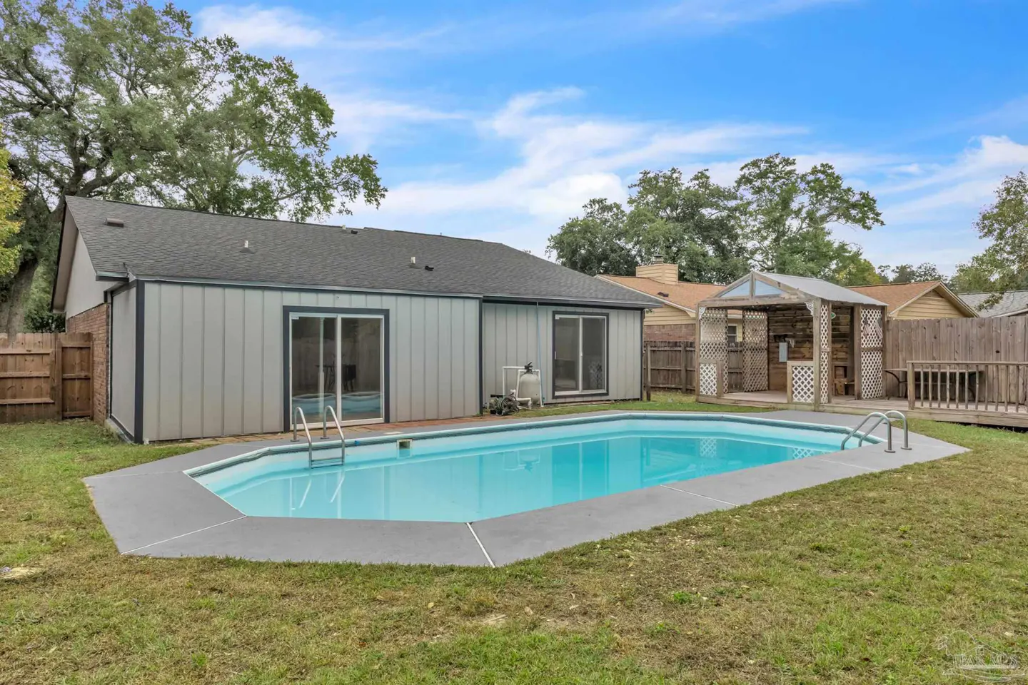 Backyard view of a light blue in-ground pool with a gray concrete border, a gray house with sliding glass doors, and a wooden gazebo.