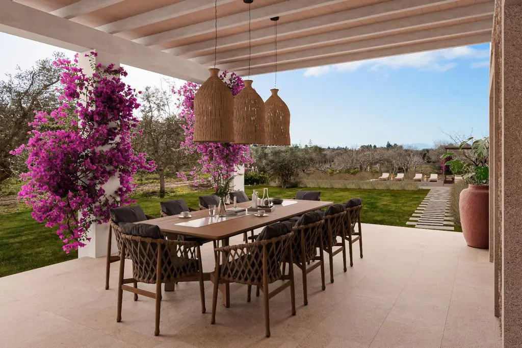 Outdoor dining area with a long wooden table, wicker chairs, and hanging lamps. Bougainvillea climbs the white columns, overlooking a green lawn and blue sky.