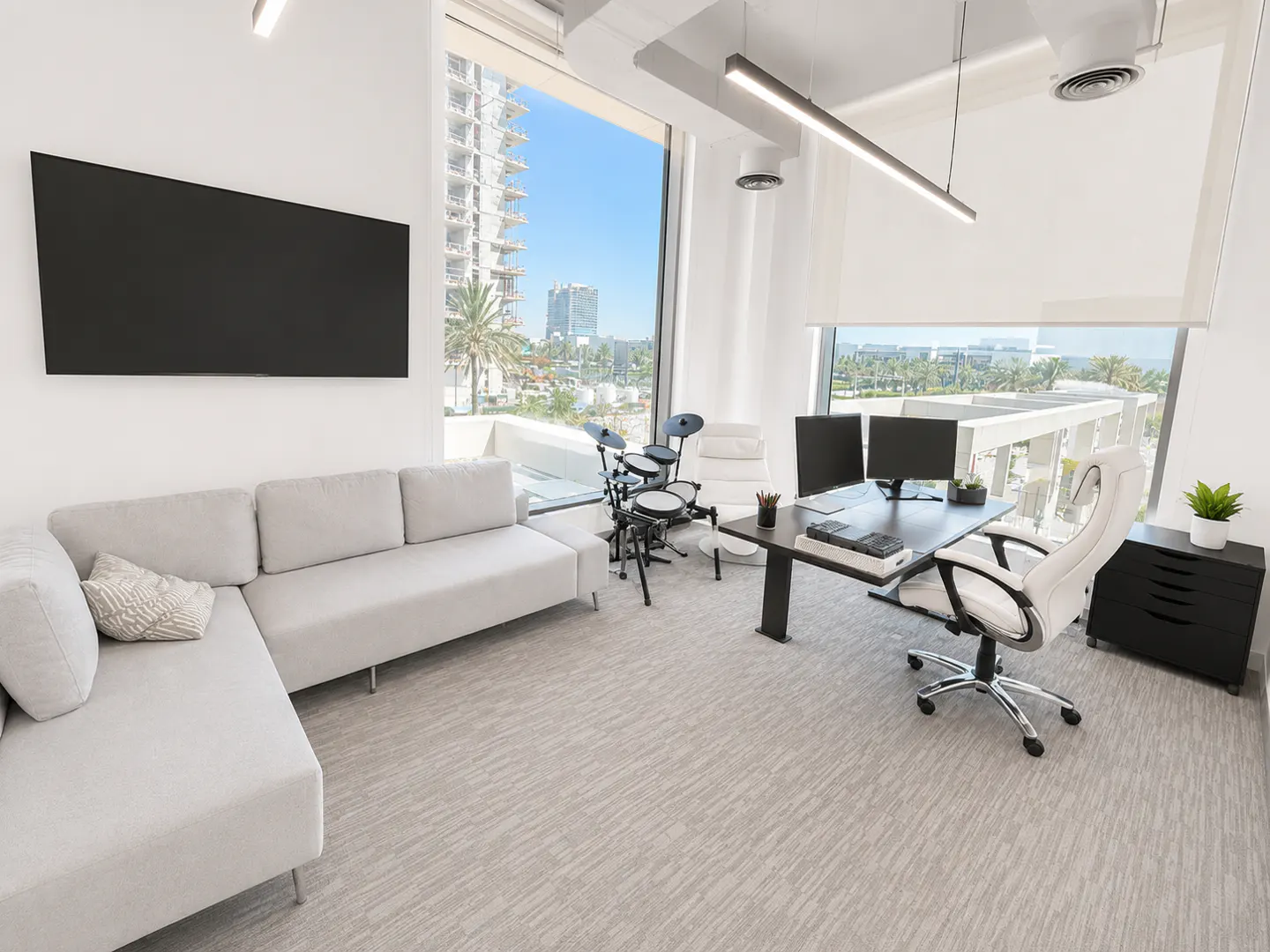 Modern office with a light gray sectional sofa, a desk with dual monitors, a white chair, and a small drum kit by the window.