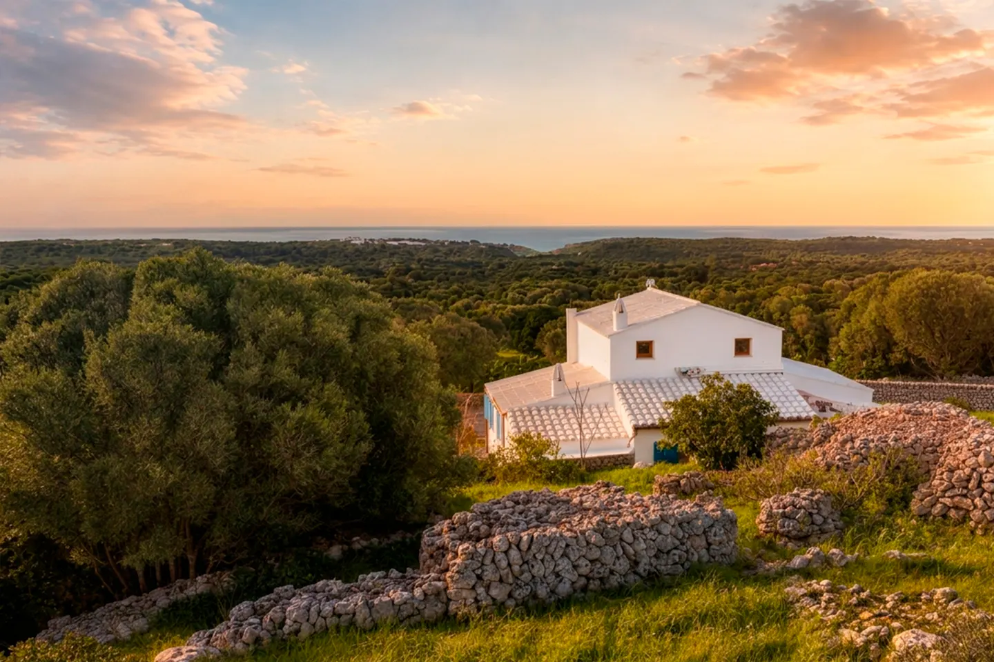 A white house with a tile roof sits on a hill overlooking a forest and the ocean at sunset.