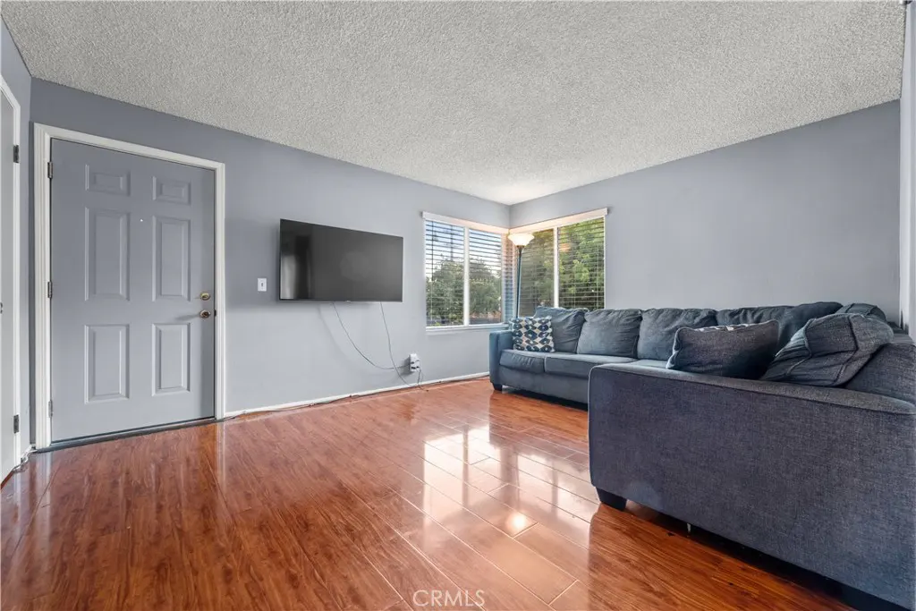 Living room with gray walls, wood floors, and a large gray sectional sofa. A flat-screen TV hangs on the wall. Natural light from a window.