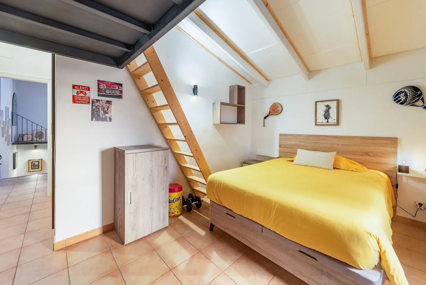 Bedroom with yellow bedding, wood headboard, and ladder to loft. Sports equipment and posters decorate the white walls.