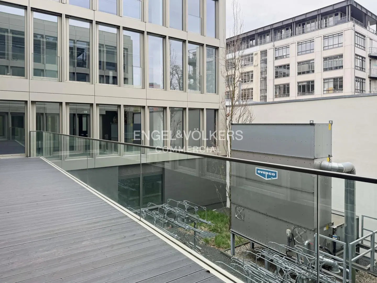 Exterior view of a modern commercial building with large windows and a gray wooden deck with glass railings. An Engel & Völkers Commercial logo is visible.