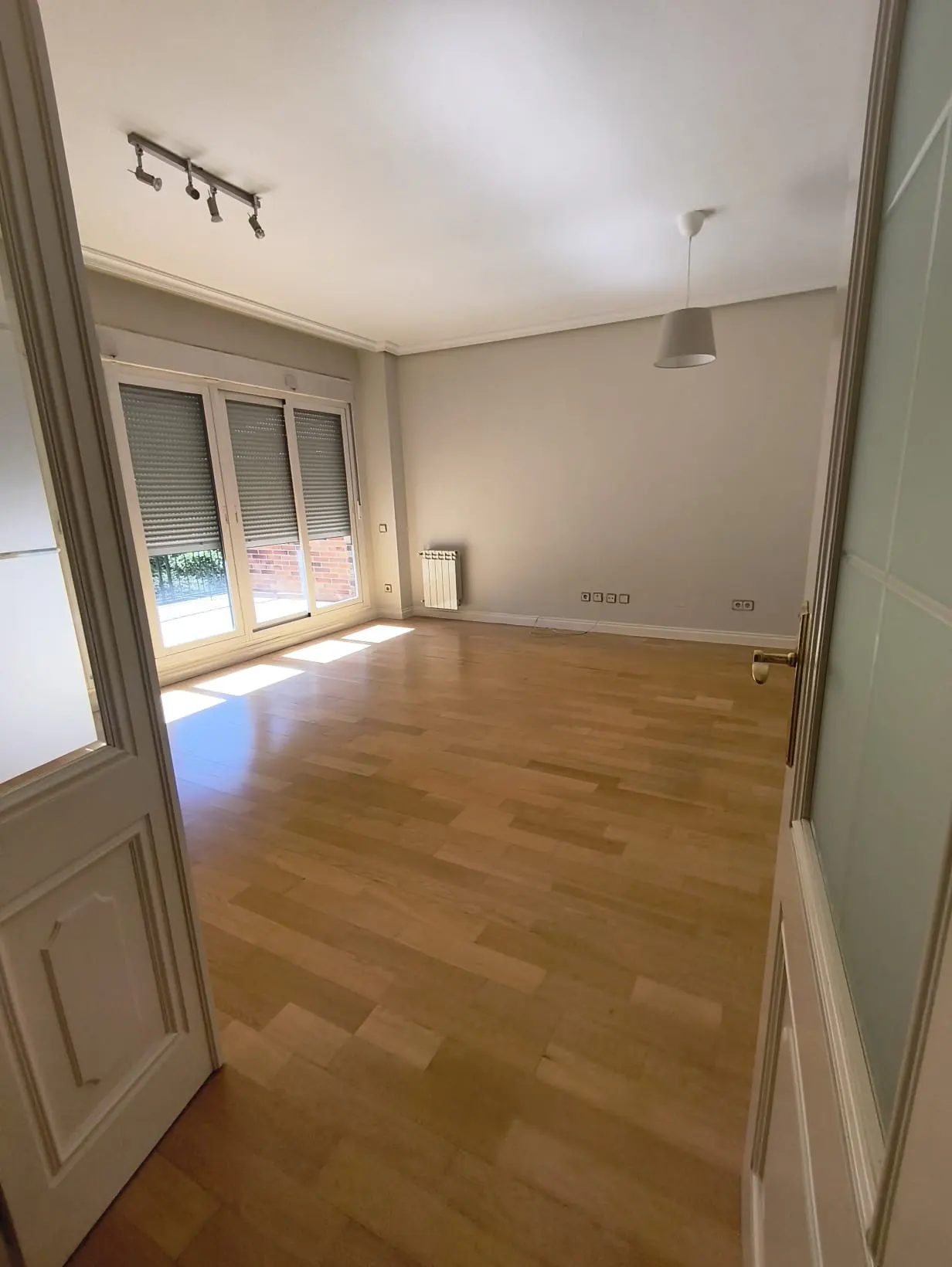 Empty living room with light wood floors, white walls, a radiator, and large windows with closed shutters.
