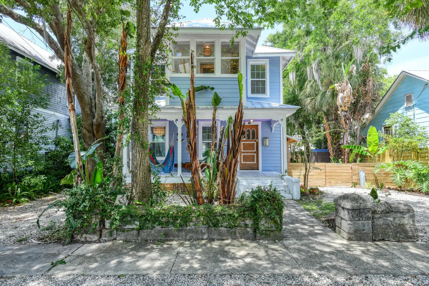 Two-story light blue house with white trim, a brown door, and a hammock on the porch. Trees and plants surround the house.
