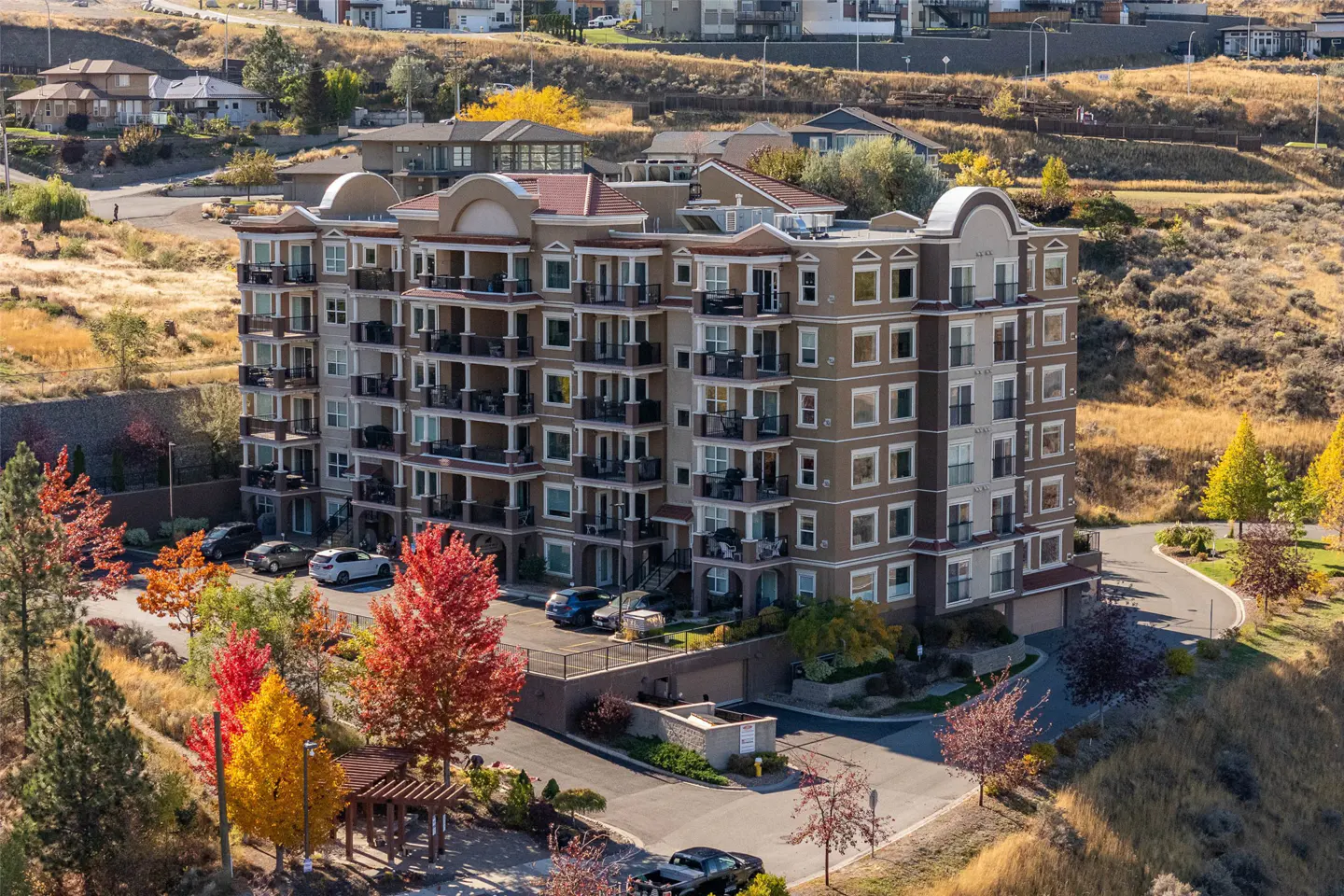 A six-story tan apartment building with balconies, surrounded by colorful autumn trees and parked cars.