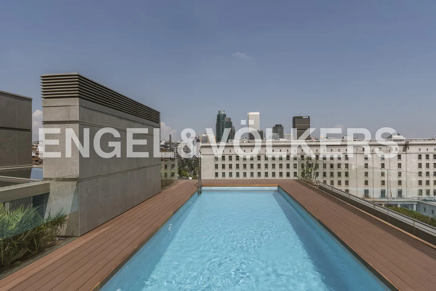 Rooftop pool with blue water and wood deck. City skyline in background under a blue sky.