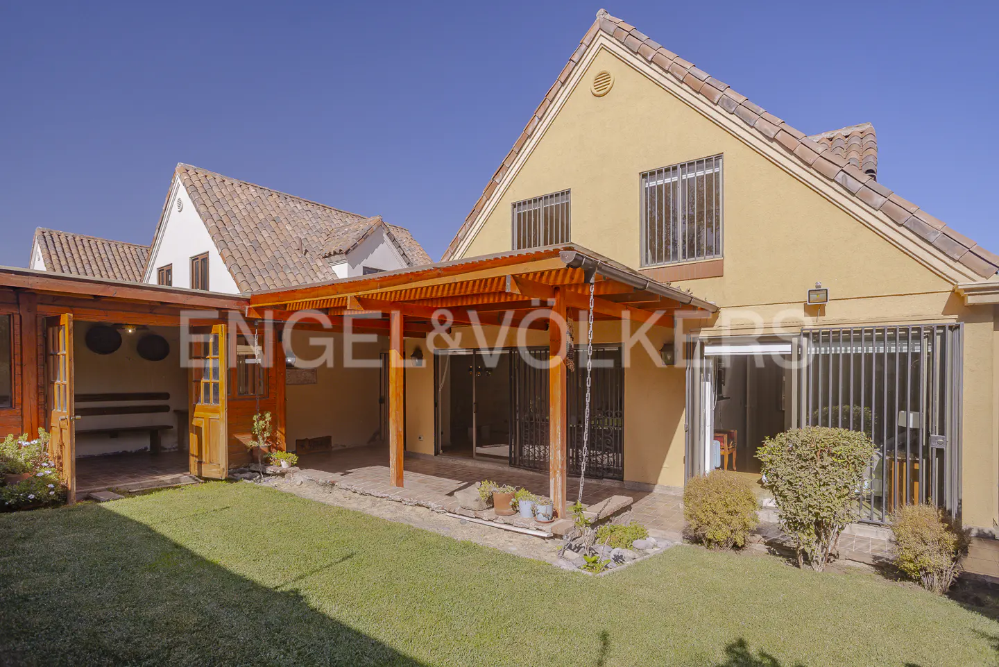 Backyard view of a two-story yellow house with a wooden pergola and green lawn under a clear blue sky.