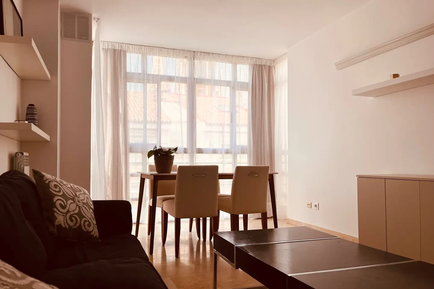 Bright living room with a dark sofa, wooden table and chairs, and a large window with sheer curtains.