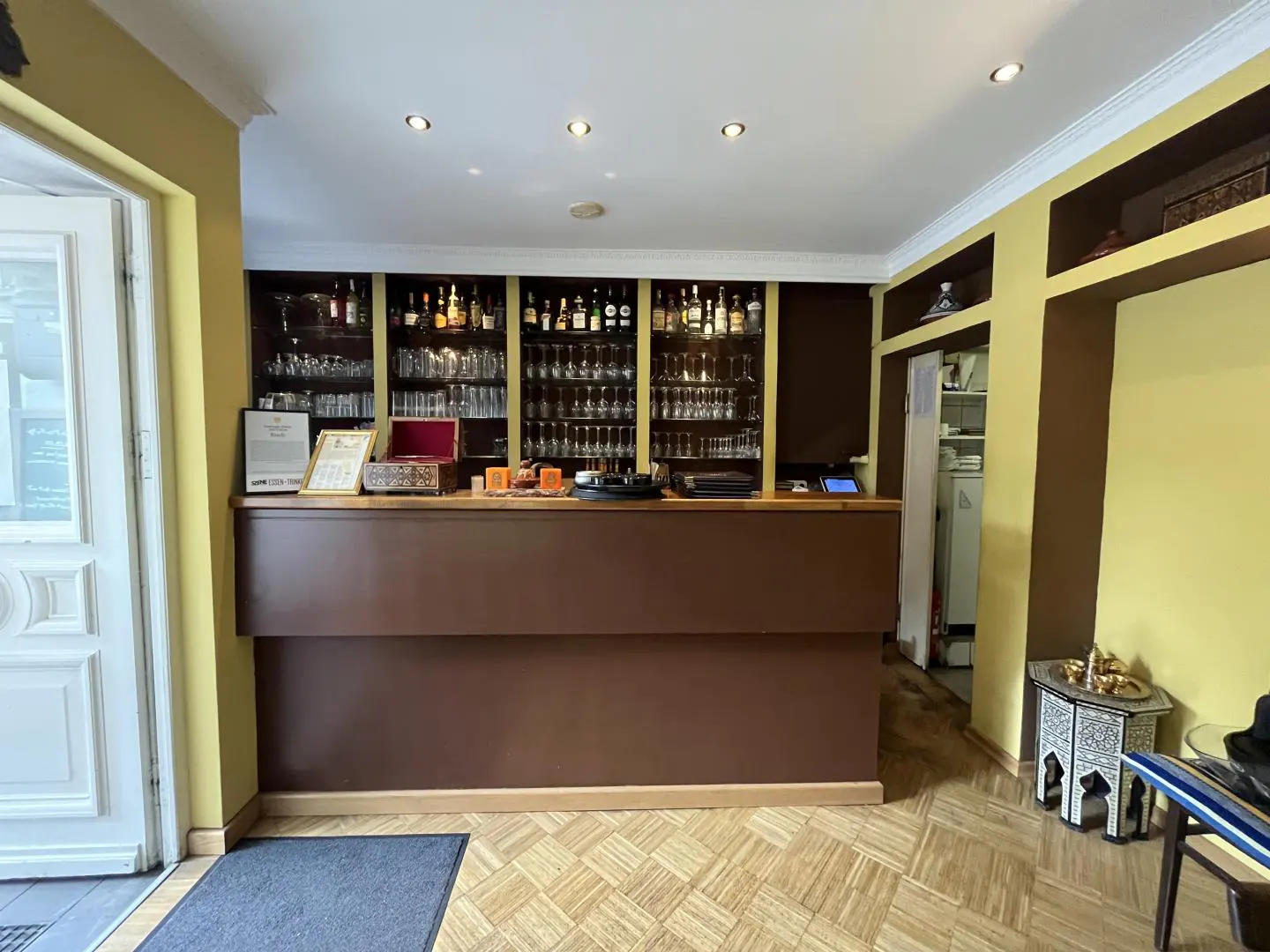 Restaurant interior with a brown bar, shelves of glasses and bottles, and yellow walls. A white door is visible on the left.