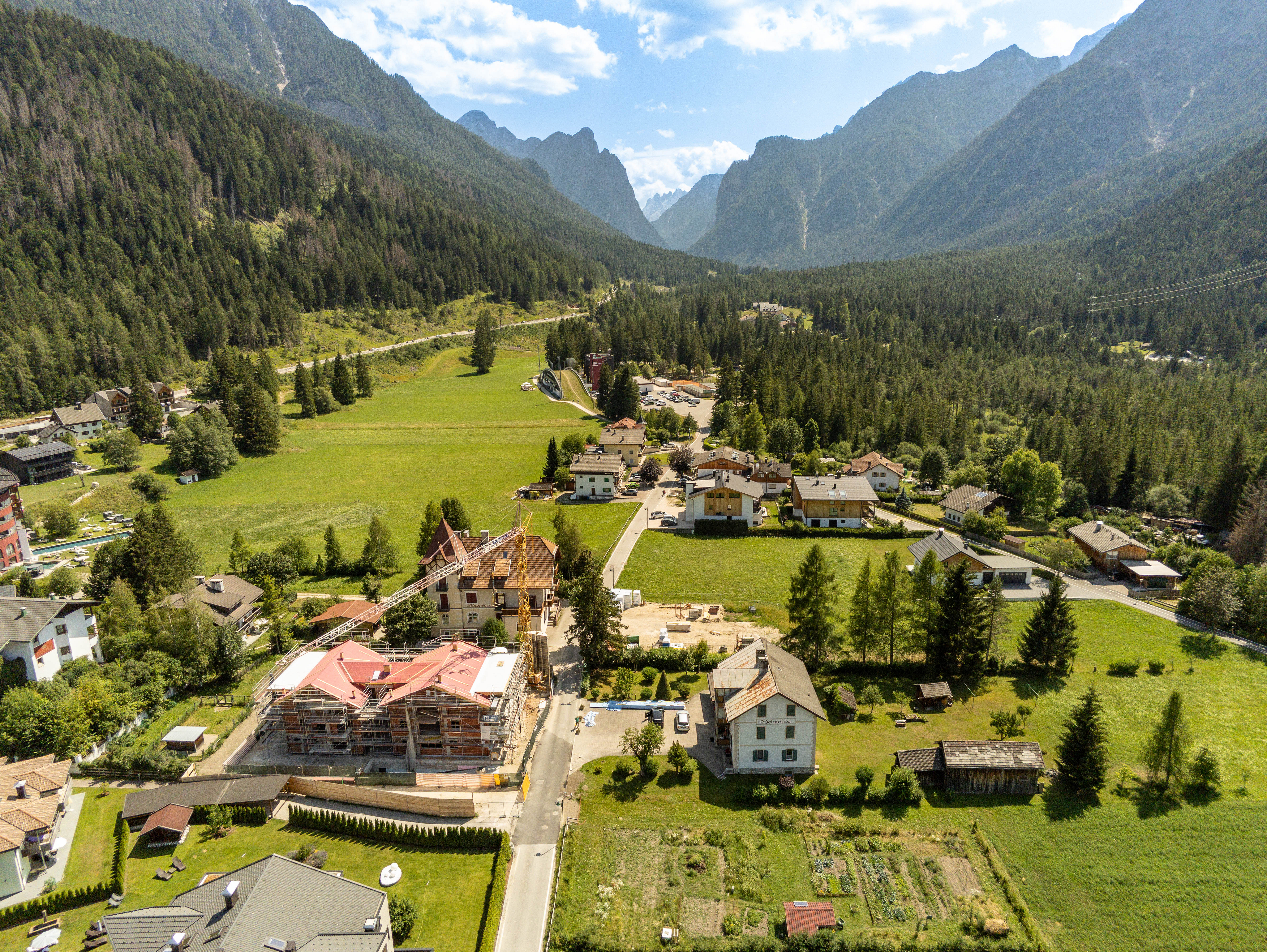Aerial view of a mountain village with a construction site, green meadows, and dense pine forests under a sunny blue sky.