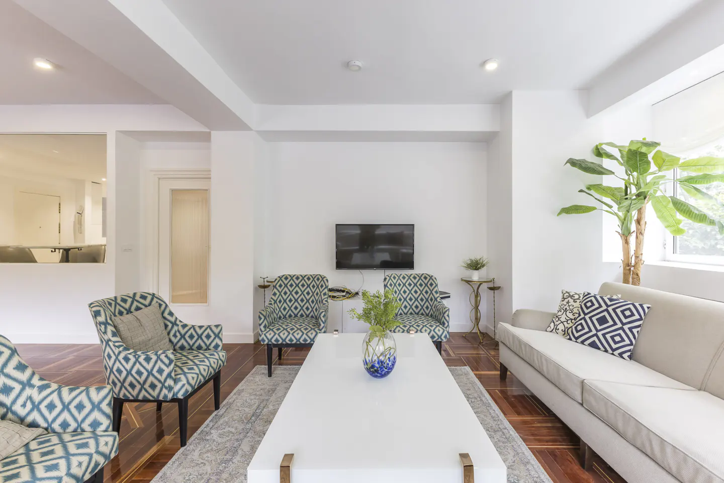 Bright living room with white walls, wood floors, and a white sofa. Patterned chairs and a large white coffee table add style. A TV hangs above.