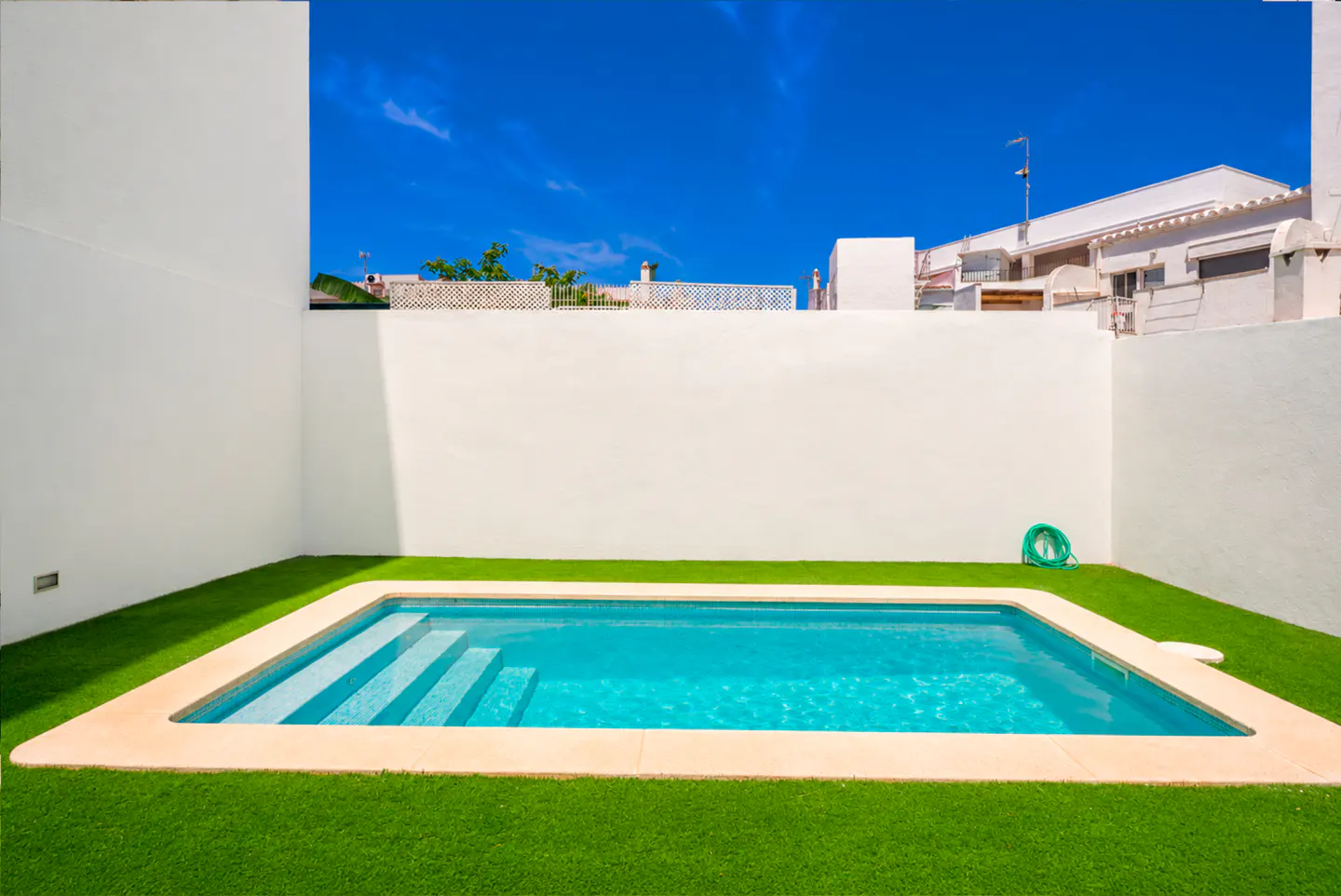 A rectangular swimming pool with turquoise water and built-in steps, set in a yard with green turf and white walls.