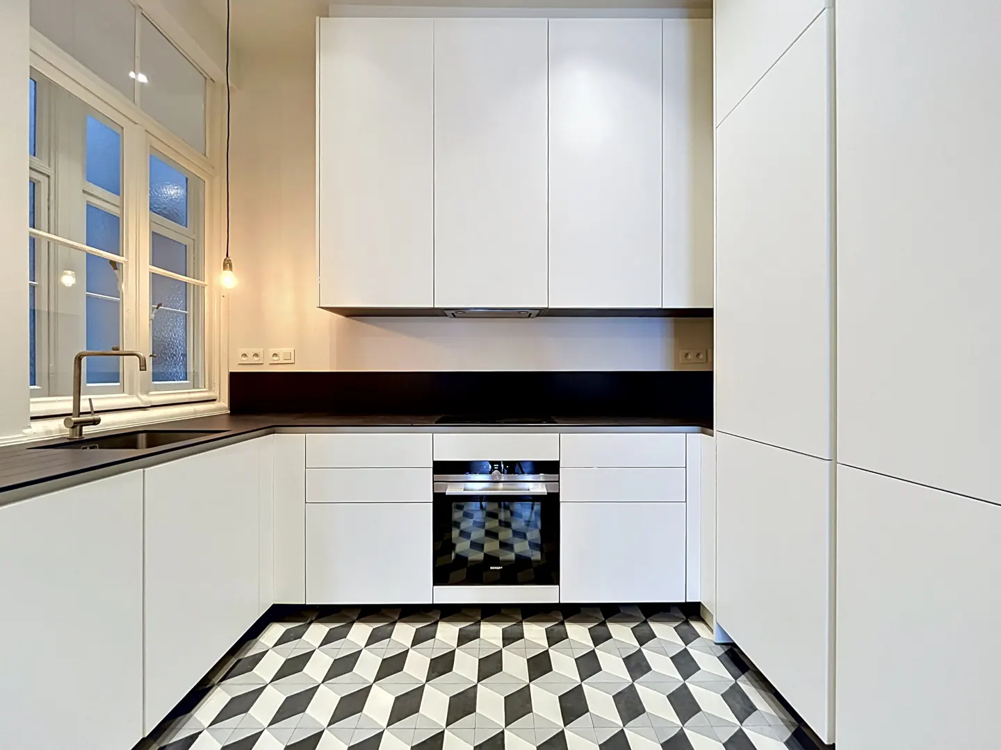 A modern kitchen with white cabinets, black countertops, and a black and white geometric tile floor. A window is on the left side of the image.