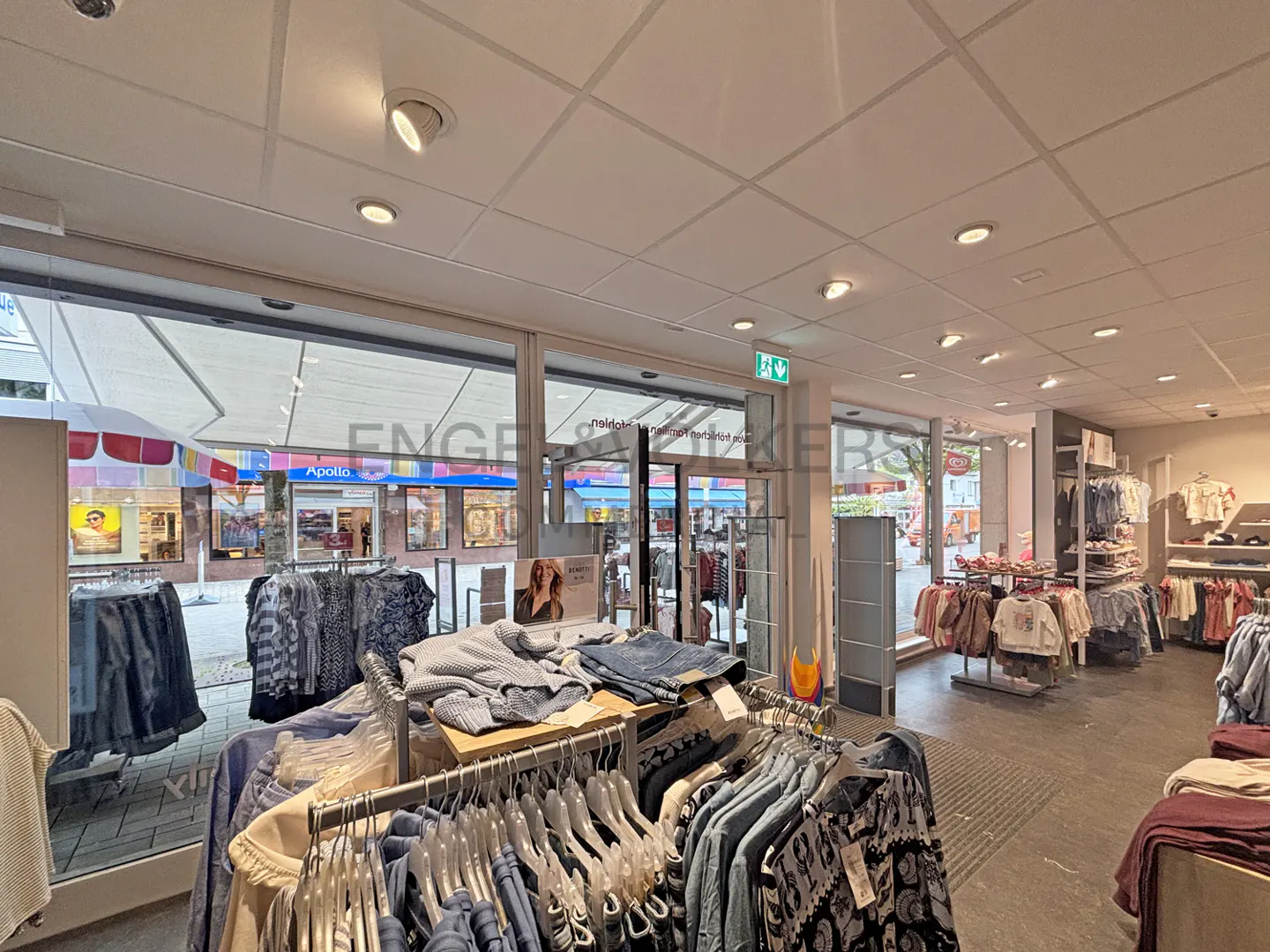 Interior view of a clothing store with racks of clothes, a gray floor, and a large window to the street.