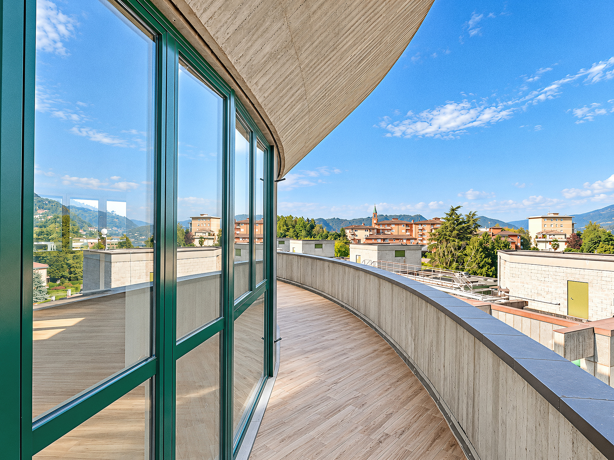 Curved balcony with wood-look flooring and concrete walls, overlooking a town with mountains under a bright blue sky. [AI-generated text]