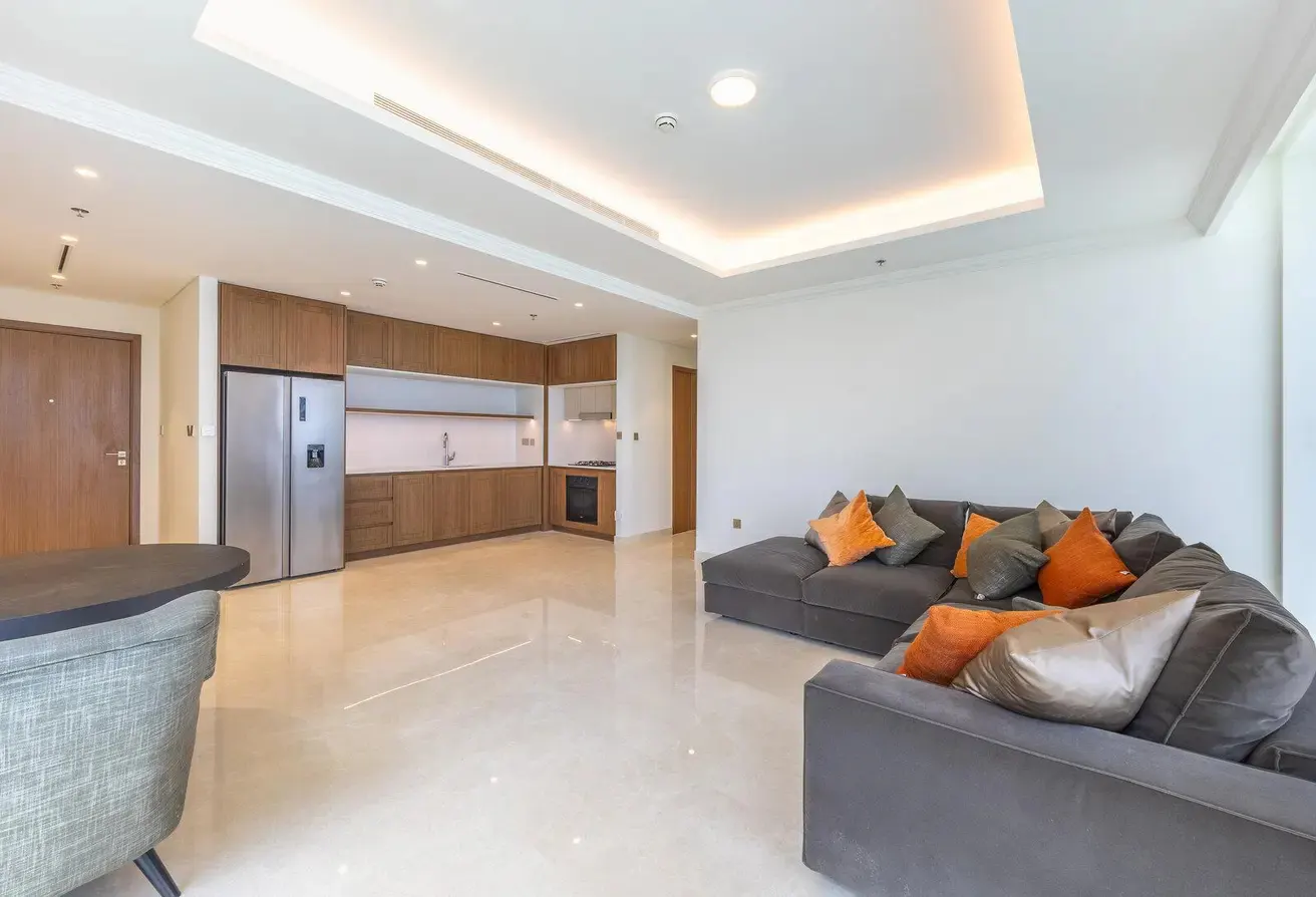 Open-concept living space with a gray sectional sofa, orange pillows, and a modern kitchen with wood cabinets.