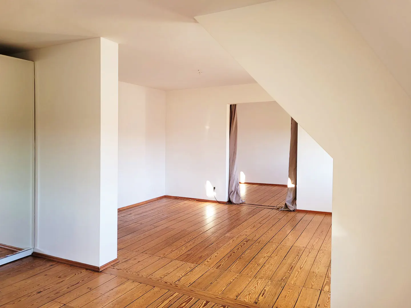 Bright attic room with wood floors, white walls, and a doorway with gray curtains.