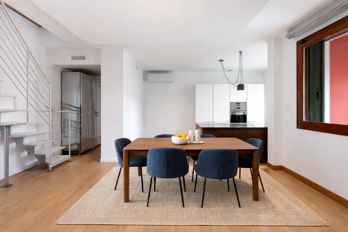 Bright, modern dining area with a wood table, blue chairs, and a jute rug. A white kitchen and metal staircase are in the background.