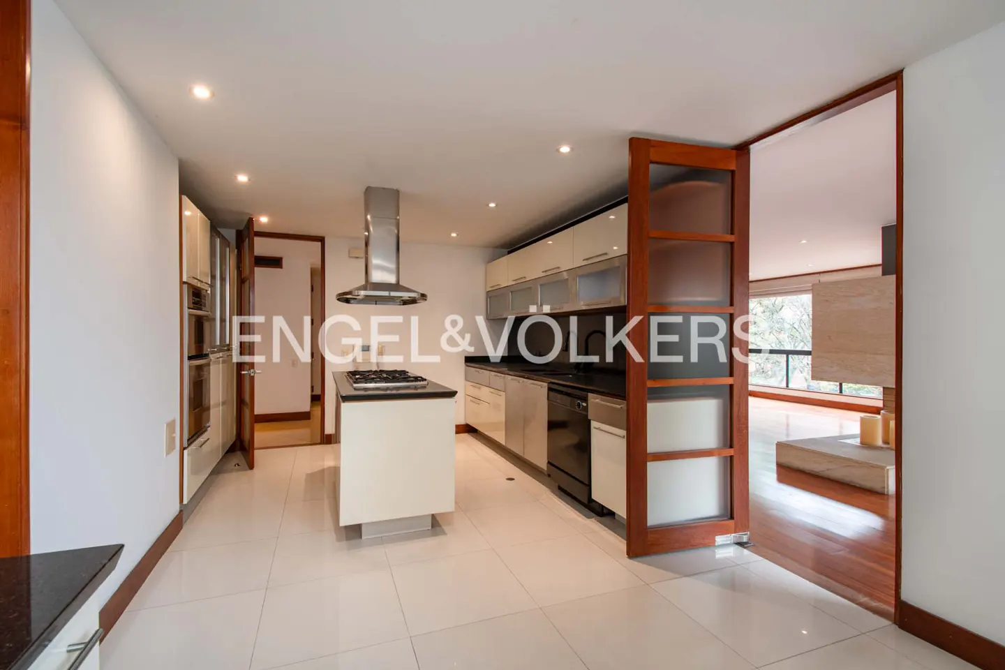 A modern kitchen with white cabinets, stainless steel appliances, and a central island with a cooktop. A doorway leads to a bright living room.
