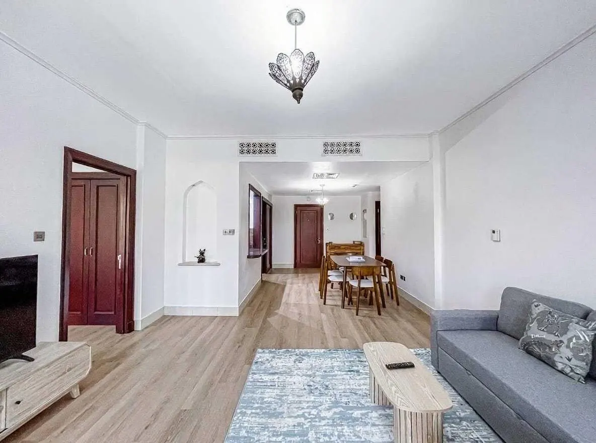 Bright living room with wood floors, a gray sofa, and a light wood coffee table with a remote. A dining table is visible in the background.