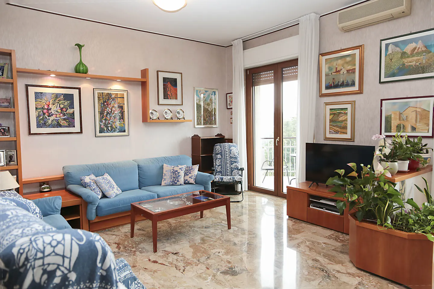 Living room with blue sofa, wood furniture, and marble floors. Paintings adorn the walls, and plants add a touch of nature. Balcony access.