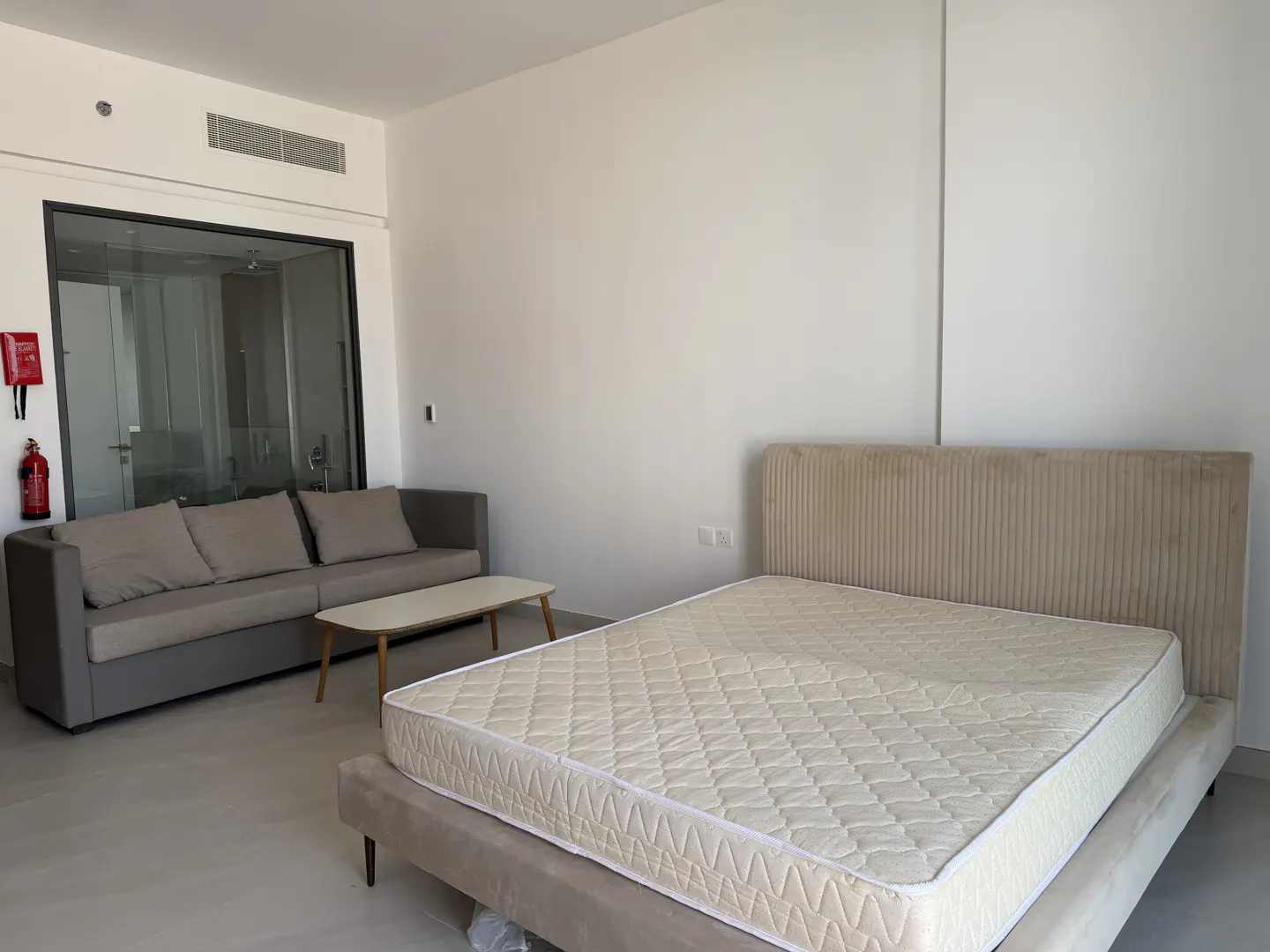 Studio apartment with a gray sofa, white coffee table, and a queen-sized bed with a beige headboard. A fire extinguisher is mounted on the wall.