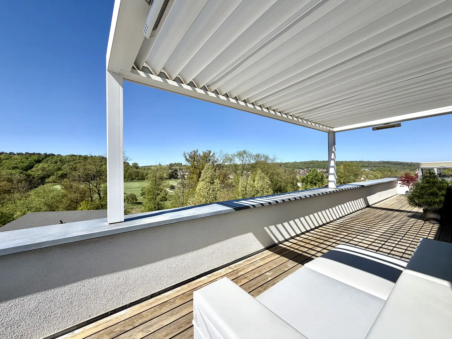 A modern white pergola shades a wooden deck with white seating, offering a scenic view of green trees and a clear blue sky.