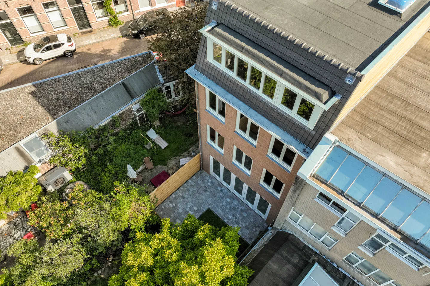 Aerial view of a brick townhouse with a paved patio, lush garden, and a modern rooftop extension with large windows.