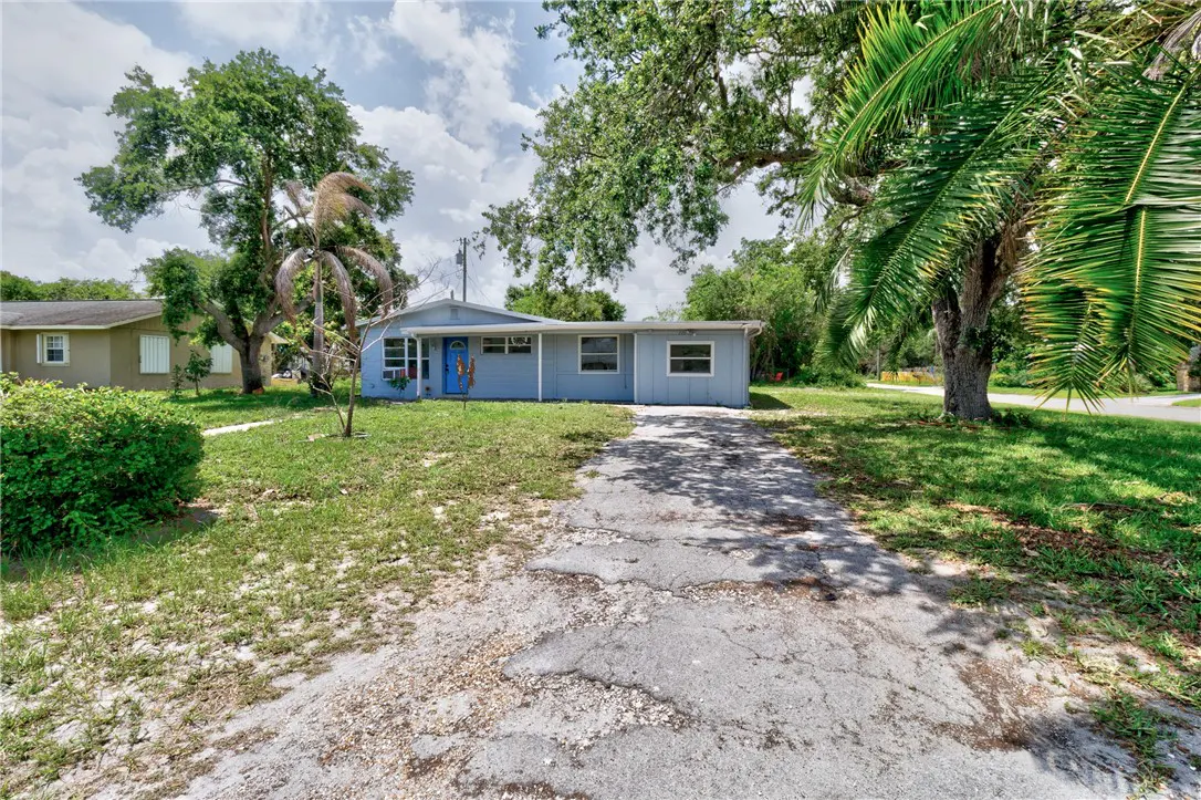 A light blue single-story house with a bright blue door and a cracked driveway. Trees and green grass surround the property.