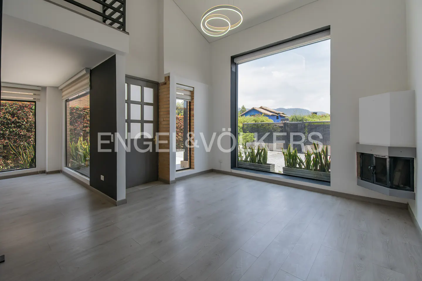 Bright, modern living room with gray wood floors, large windows, and a fireplace. Outside view of greenery and a blue-roofed house.