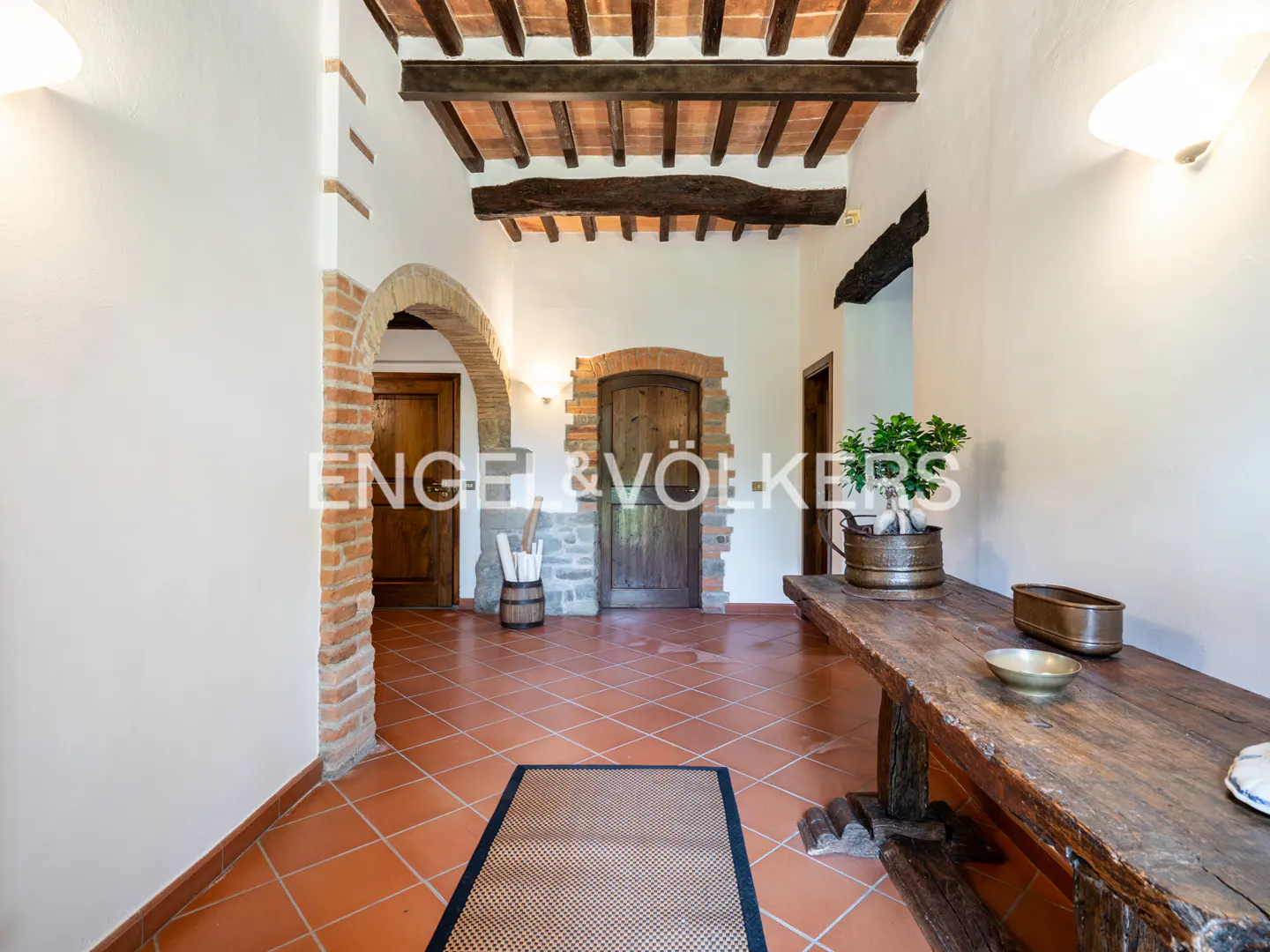 Interior view of a Tuscan-style foyer with terracotta tile floor, wooden doors, and exposed beam ceiling. A rustic wooden table sits to the right.
