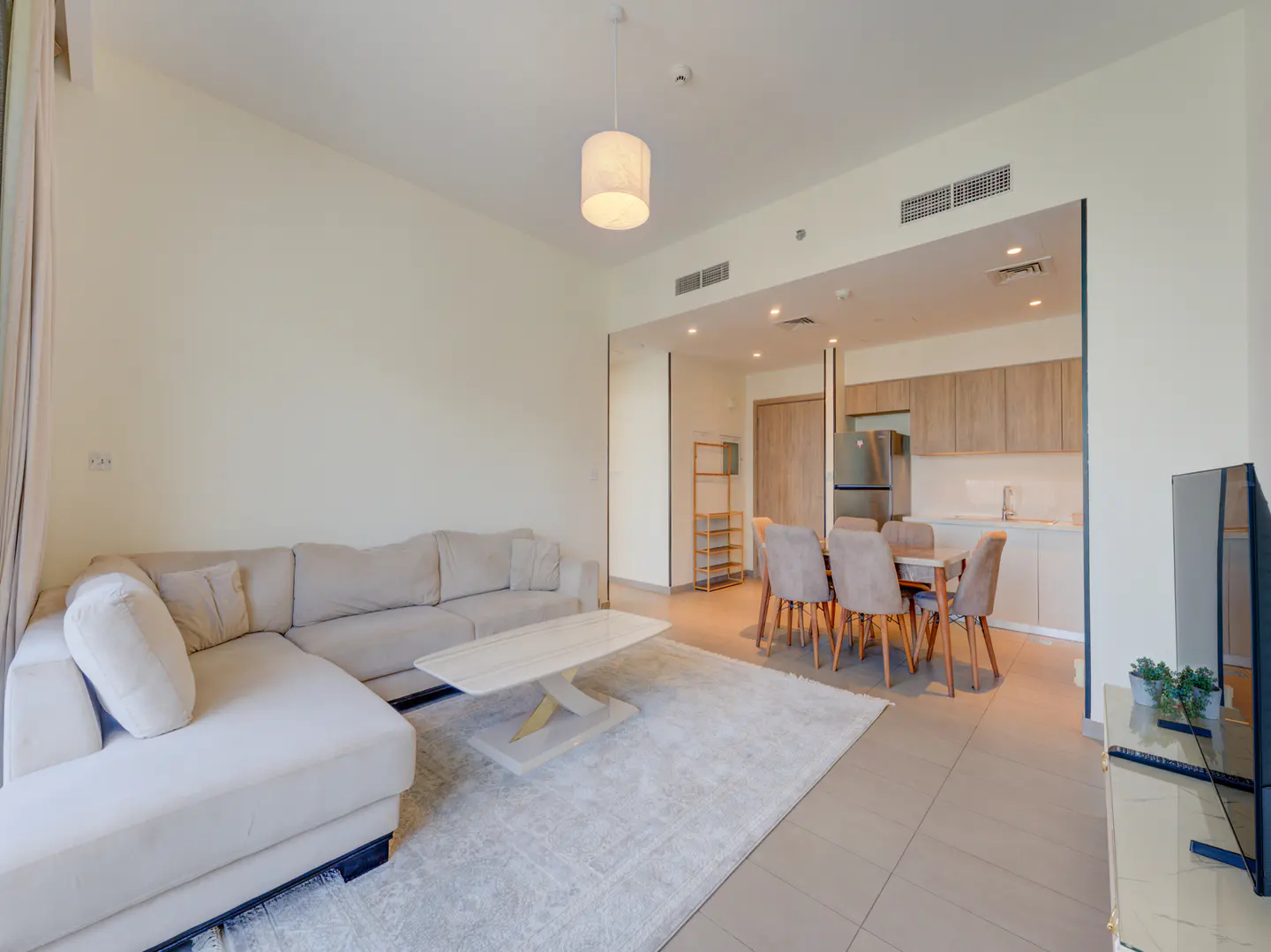 Bright apartment interior with a beige sectional sofa, marble coffee table, dining set, and kitchen in the background.