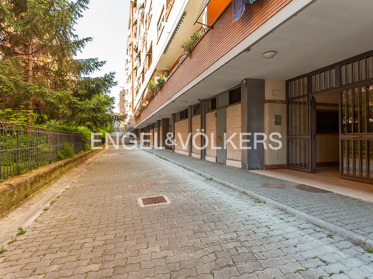 Exterior view of a multi-story apartment building with a brick and concrete facade, a paved walkway, and green trees.