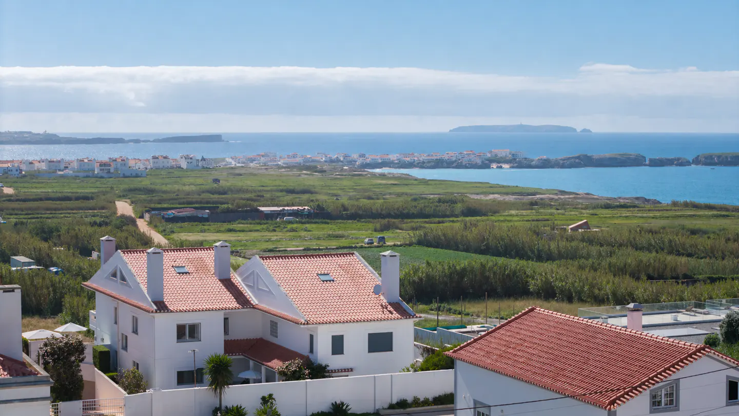 Scenic view of white houses with red tile roofs, green fields, and the blue ocean under a bright sky.